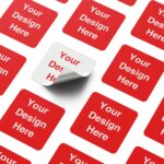 Red square sticker mockup sheet with 'Your Design Here' placeholders printed across the sheet, with one sticker being peeled up to show the adhesive backing; ideal for showcasing custom sticker printing templates.